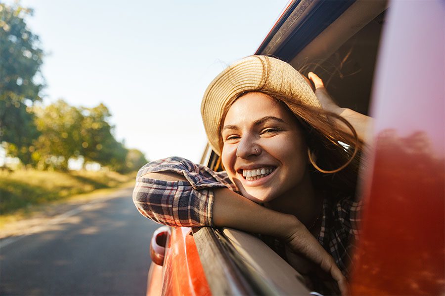 About Our Agency - View of a Cheerful Young Woman Sticking Her Head Out the Window from the Passenger Seat in a Car Enjoying the Fresh Air and Countryside Views