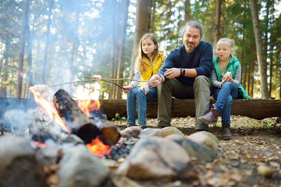 Blog - Father and His Two Daughters Sitting on a Log in the Woods During a Camping Trip Having Fun Toasting Marshmellows